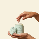 A person's hands holding an open jar of massage body butter with cream being dispensed, set against a solid light beige background.
