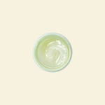 A top-down view of a small round container filled with a pale yellow-green cream or gel, centered on a solid light beige background.