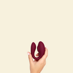 Hand holding a plum colored blended stimulation vibrator on a beige background