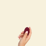 Hand holding the remote for the plum colored blended stimulation vibrator on a beige background